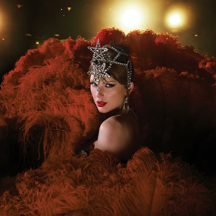 Taylor Swift in elaborate showgirl costume with red feathers and jeweled headpiece in dramatic lighting.