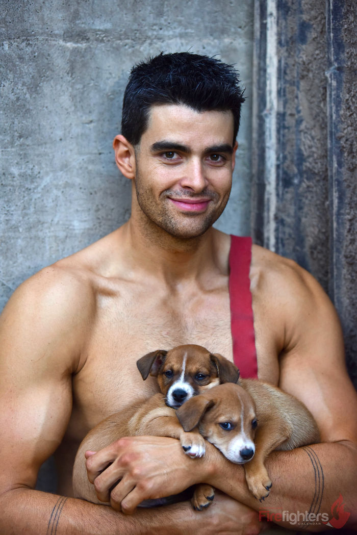 Muscular firefighter holding two adorable puppies, showcasing heroic firefighters and adorable dogs from the Australian calendar.