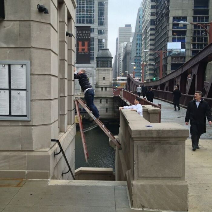 Man precariously leaning on a ladder over a river with others nearby on a city bridge, a risky image that might need an exorcist.