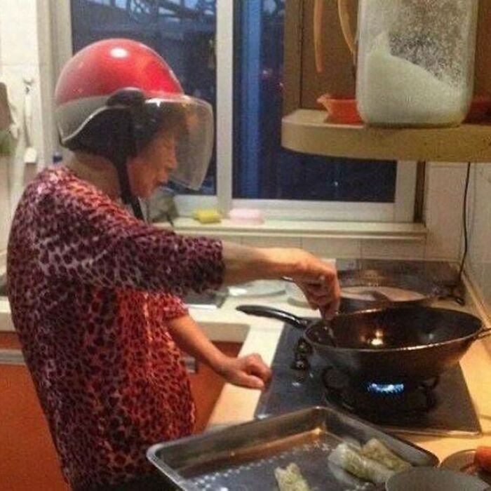 Elderly woman wearing a red helmet while cooking on a stove in a kitchen, a funny image needing exorcist humor.
