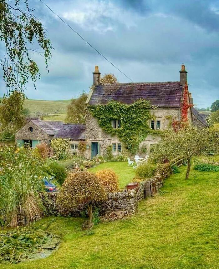 Stone dream cottage covered in ivy with a vibrant garden and lush green lawn on a cloudy day.