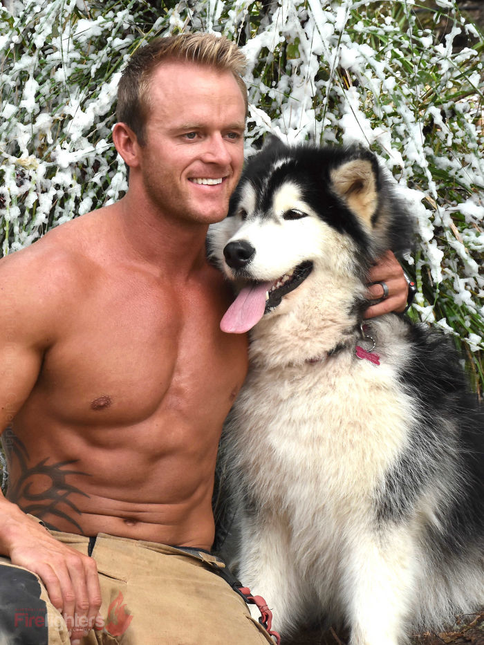 Shirtless heroic firefighter posing outdoors with an adorable fluffy dog with tongue out against a snowy branch background