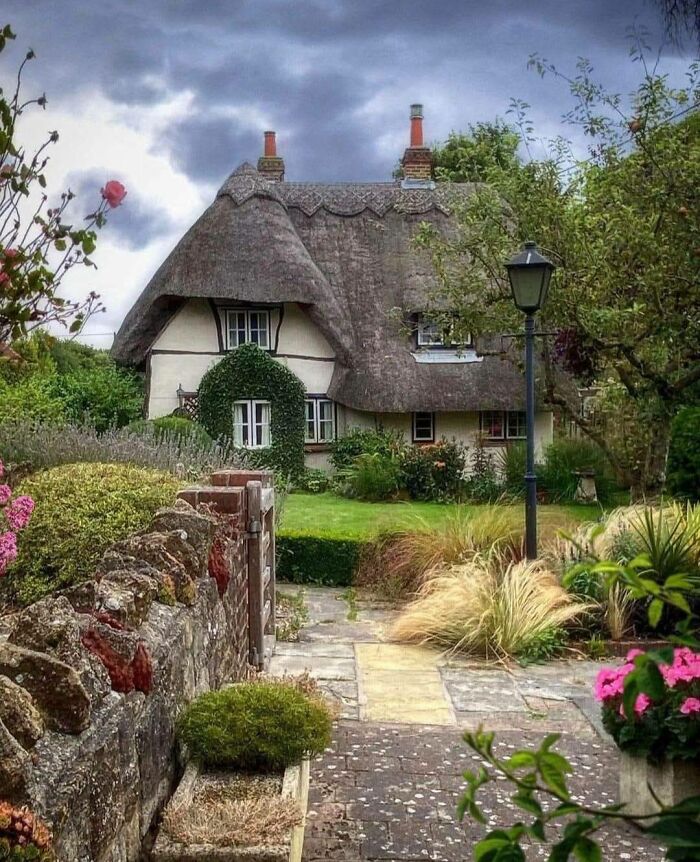 Idyllic dream cottage with thatched roof surrounded by lush gardens and stone pathways under a moody sky.