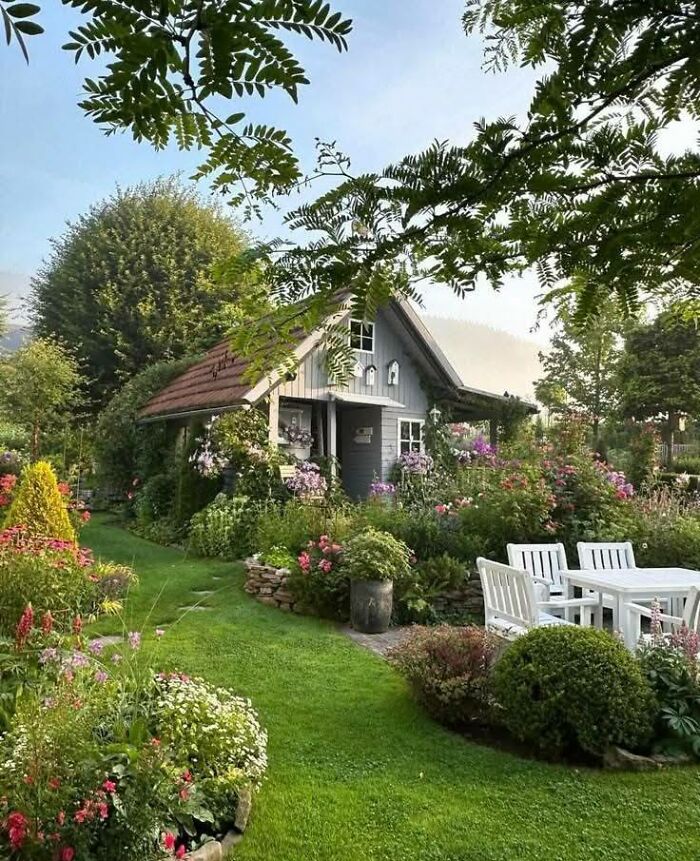 Idyllic dream cottage surrounded by lush gardens with blooming flowers and outdoor white furniture on green lawn.