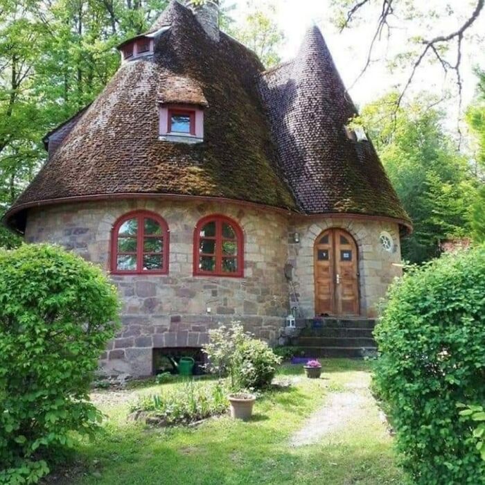 Cozy dream cottage with unique curved roof surrounded by lush green gardens and vibrant plants in a peaceful setting.