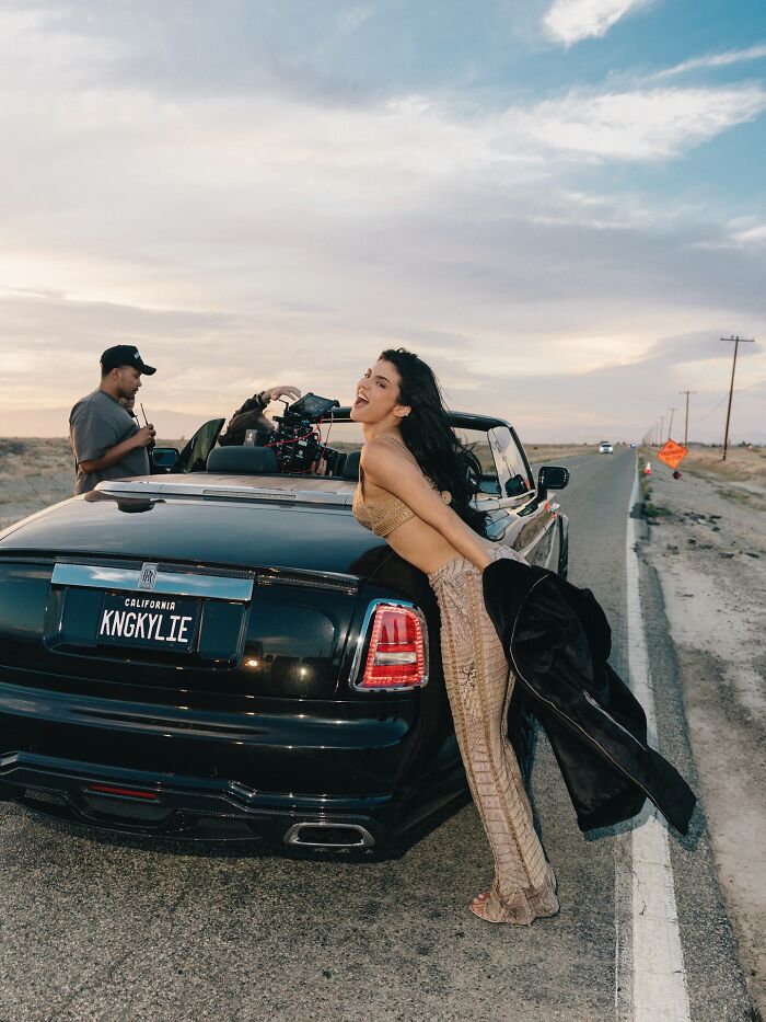 Woman posing by a luxury car with a personalized license plate KNGKYLIE during a roadside video shoot amid fake arrest controversy.