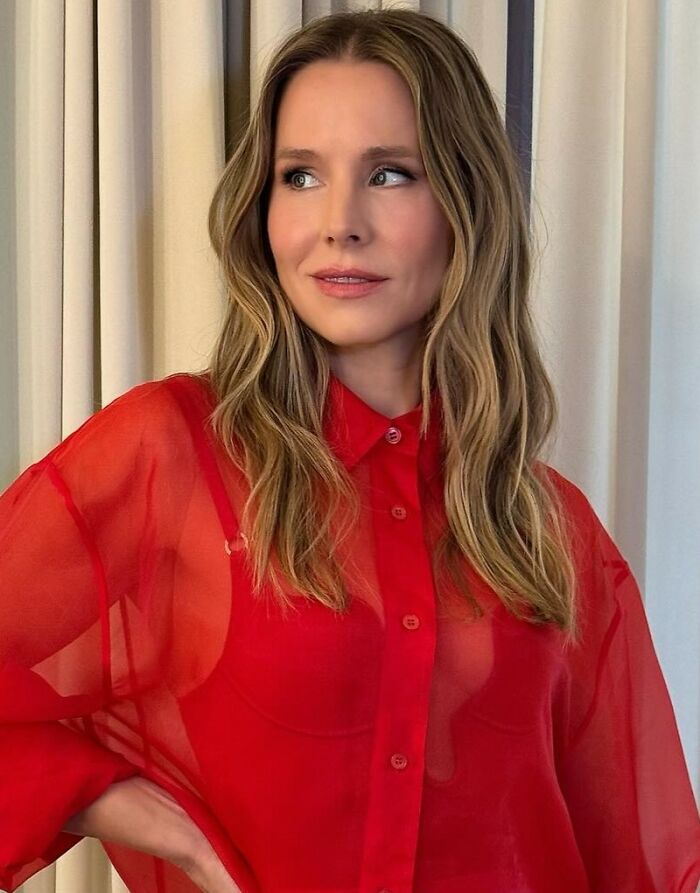 Kristen Bell in a red sheer blouse, looking to the side with a neutral expression against a cream curtain backdrop.