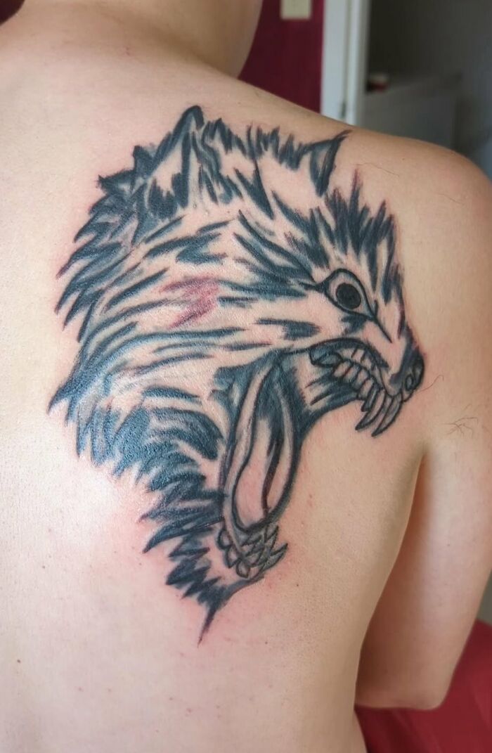 Large black and gray wolf head tattoo with open mouth and sharp teeth on a person's upper back, unusual tattoos.