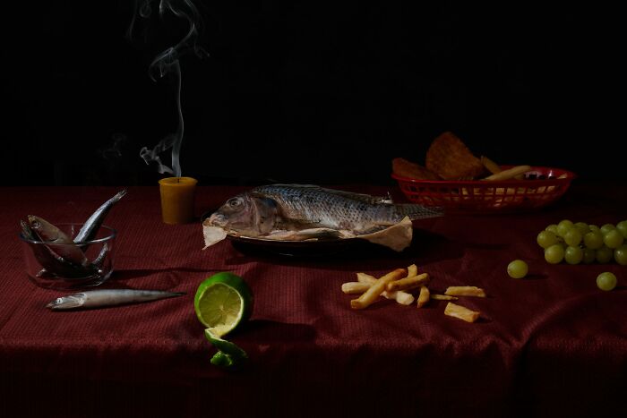Still life featuring a chicken, fried chicken sandwich, raw poultry, garlic, grapes, and a lit candle in dark setting.