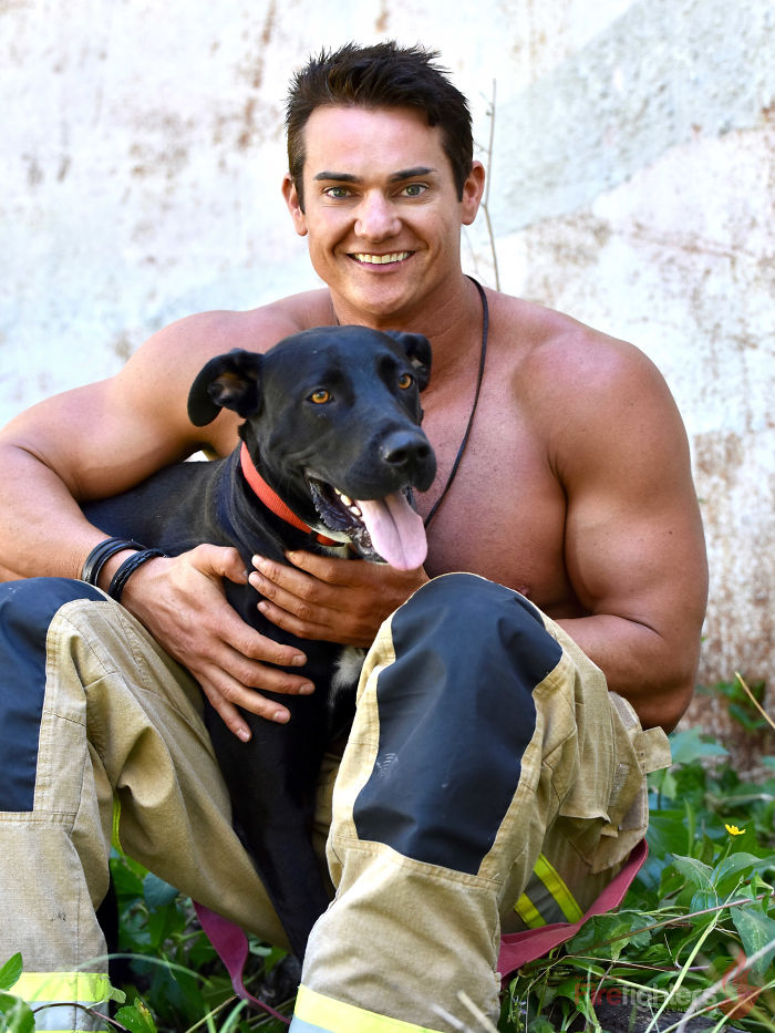 Muscular firefighter sitting outdoors with adorable black dog wearing a red collar in Australian calendar photoshoot.