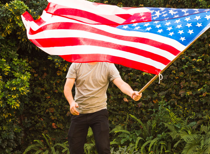 Man holding large American flag outdoors with foliage background, symbolizing refusal to attend wedding off American soil
