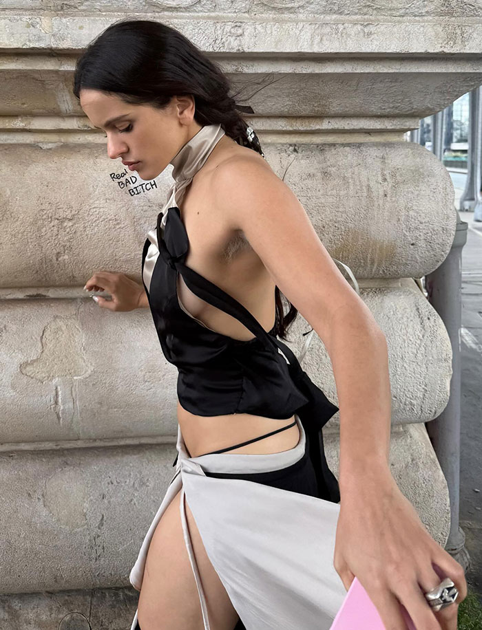 Pop star showing dyed armpit hair at Paris Fashion Week, wearing black and white outfit against stone backdrop.