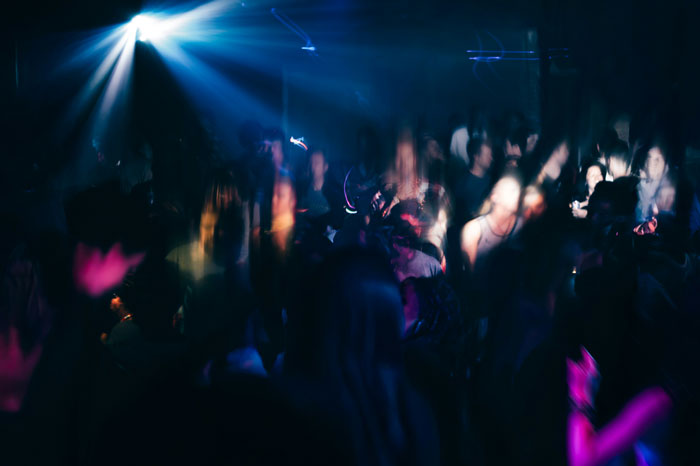Crowded dark room with blurred people and bright spotlight, capturing chaotic moments in wild we need to leave right now stories.
