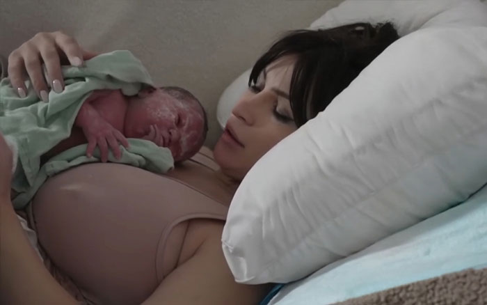 Woman giving birth on Twitch livestream holding newborn baby while resting on a bed with white pillow nearby.