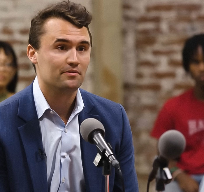 Man in blue blazer speaking into microphone during discussion about Charlie Kirk homework assignment controversy.