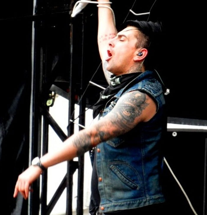Tattooed man in a sleeveless denim vest raising his arm during a live performance on stage, expressing emotion.