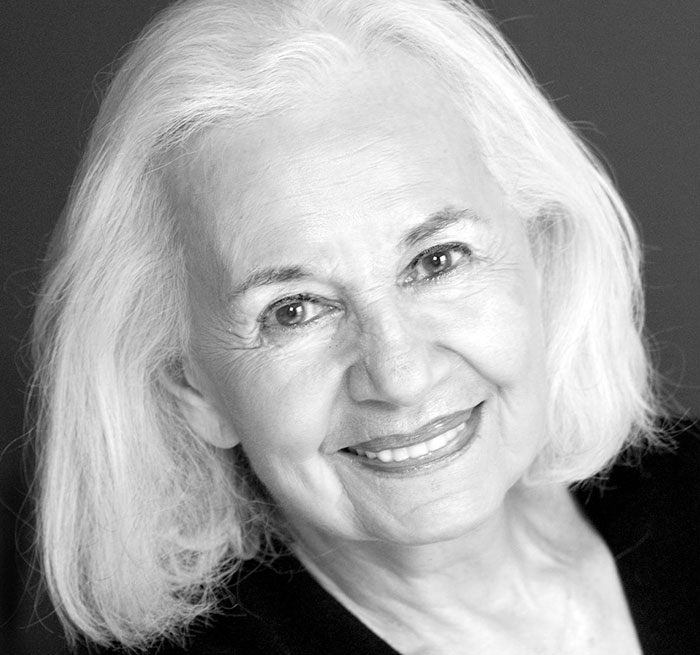 Holocaust survivor Ruth Posner smiling softly, with white hair and wearing a black top against a dark background.