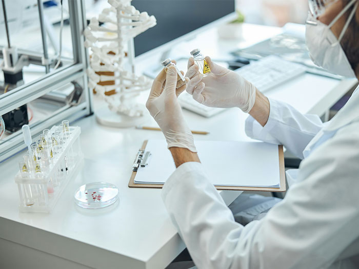 Doctor in a lab wearing gloves and mask, examining vials while reviewing notes on a clipboard next to a skeleton model.