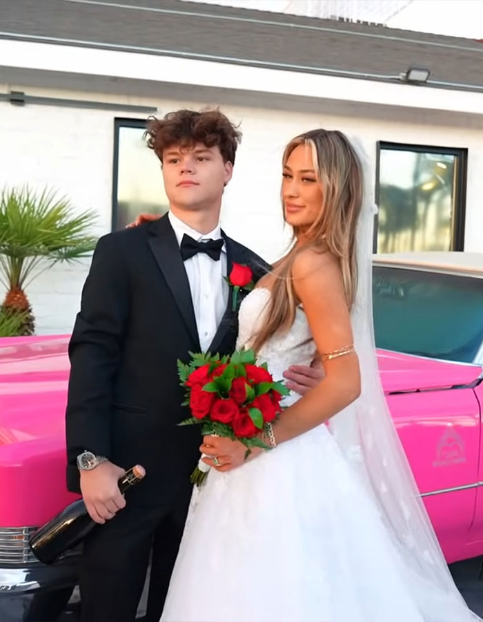 Adult star in wedding dress with groom in tuxedo holding bouquet and bottle, posing outdoors near a vintage pink car.