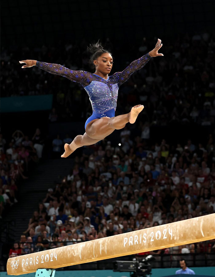 Simone Biles performing a leap on the balance beam at Paris 2024 Olympic Games with a large audience in the background.