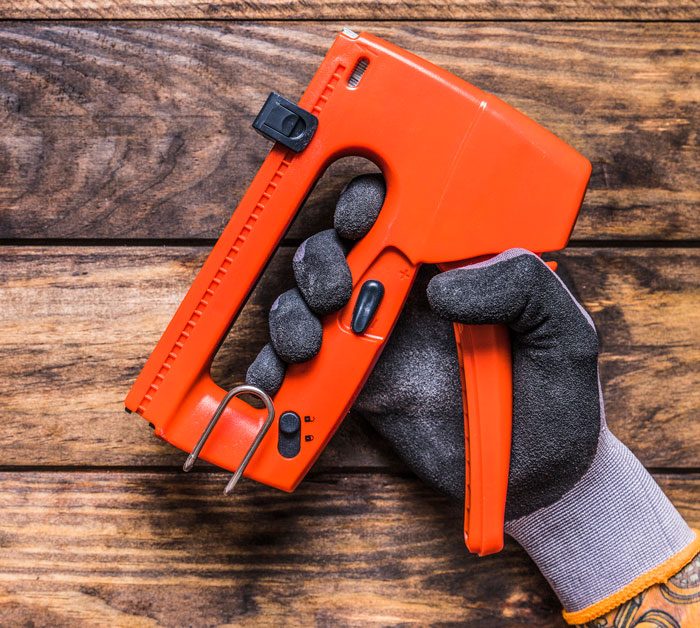 Hand in work glove holding an orange staple gun over a wooden surface, illustrating worst employees tools.
