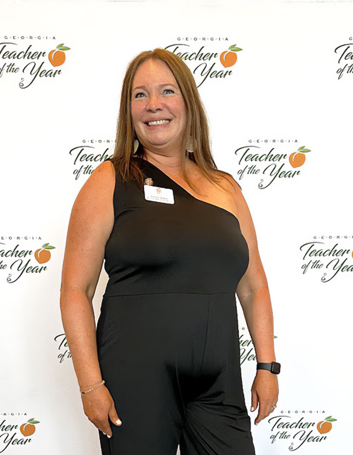 Woman in a black one-shoulder outfit smiling at a Teacher of the Year event with logo backdrop in Georgia