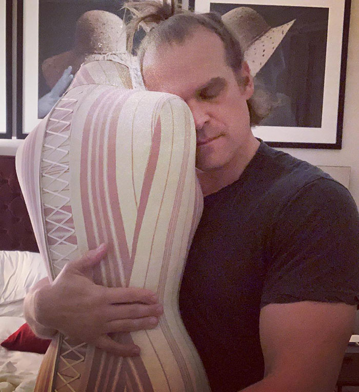 David Harbour embracing a striped mannequin, showing a calm expression in an indoor setting.
