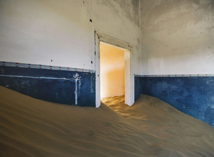 Abandoned home interior half-buried in desert sand showing surreal sand castles swallowing the structure.