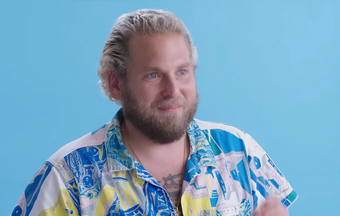 Jonah Hill showing major transformation with beard and colorful shirt against a blue background in a casual setting