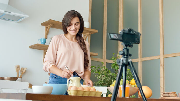 Young woman demonstrating life hacks in kitchen while recording video with camera on tripod, sharing genius tips online.