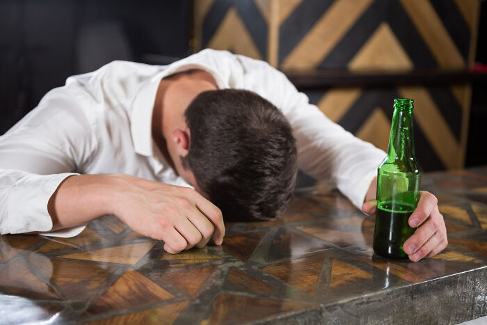 Man in white shirt with head down on bar table holding green beer bottle, illustrating restaurant staff tales about unpaid bills.