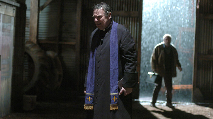 Man dressed as a priest stands in a dimly lit room with another man approaching, from a movie based on true stories.