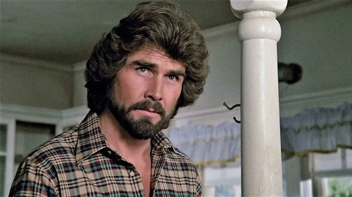 Man with beard and long hair wearing a plaid shirt in a vintage home setting, representing movies based on true stories.