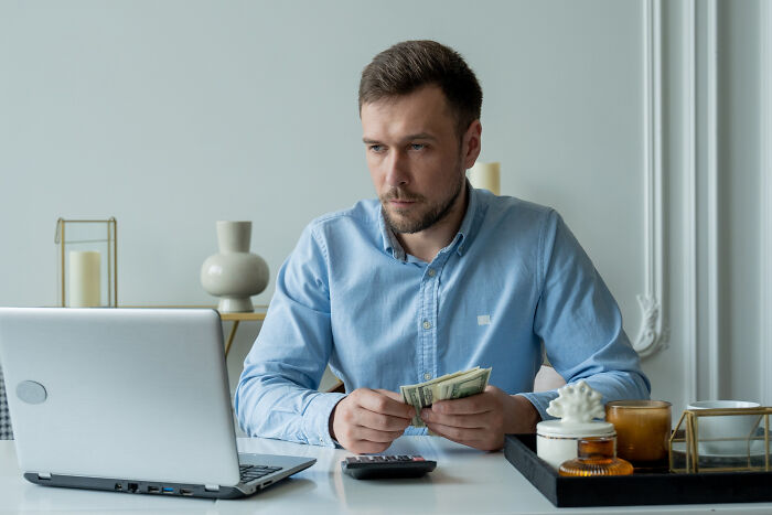Man in a blue shirt sitting at a desk with laptop and calculator, counting money and thinking about life hacks.