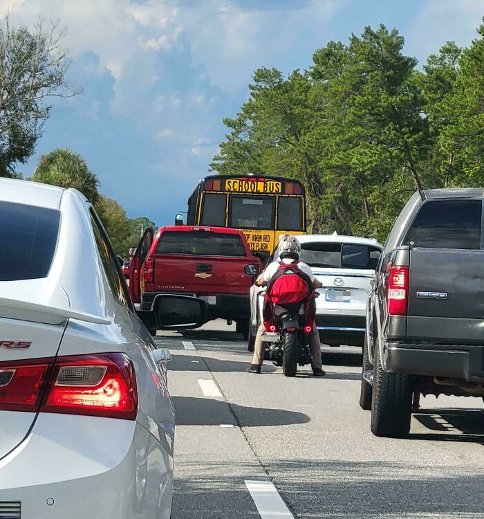 Motorcyclist squeezing between cars and a school bus in traffic, showing people allergic to acting like sensible adults.