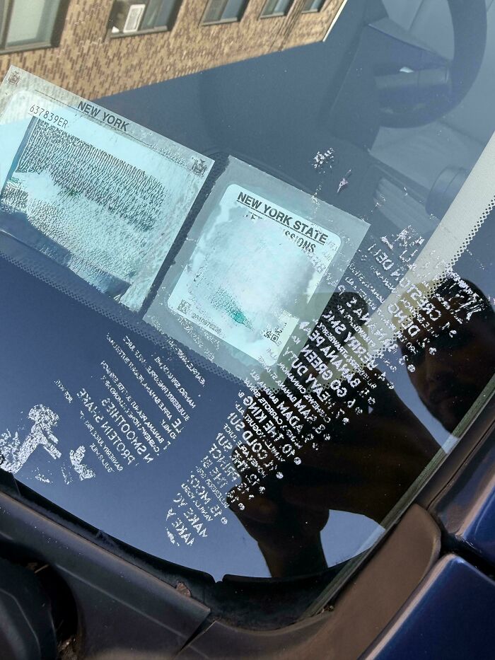 Car windshield with old New York State inspection stickers, showing wear and reflections of building and person taking photo.
