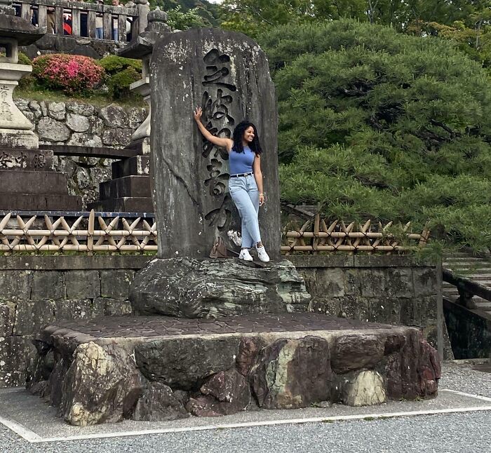 Young woman posing playfully on a stone monument in a garden, showing a carefree attitude from people allergic to acting sensible adults