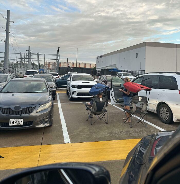Two people sitting in folding chairs with umbrellas open in a parking lot, showing people allergic to acting like sensible adults.
