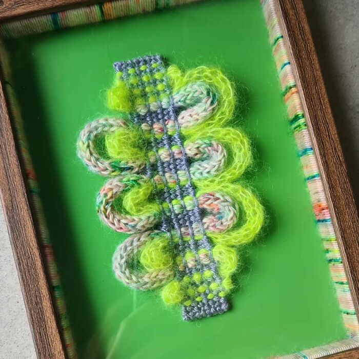 Colorful textile creation with vibrant yarn loops and intricate weaving displayed in a wooden frame on a green background.
