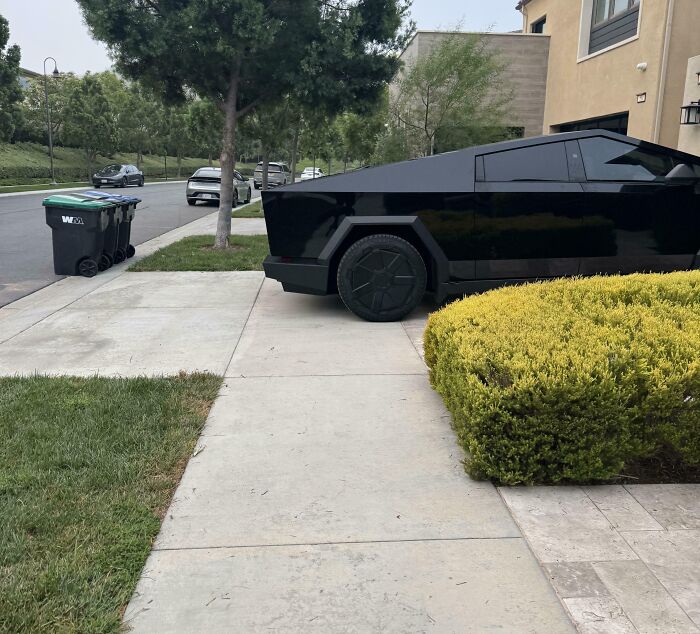 Black futuristic vehicle parked blocking a sidewalk in a residential area, showing people apparently allergic to acting like sensible adults.