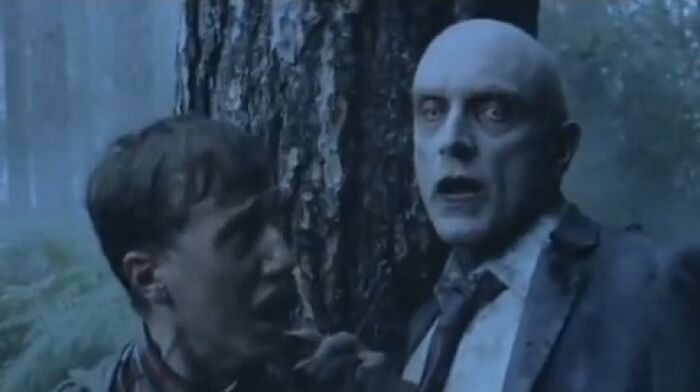 Two men in a dark forest, one with pale makeup and haunting eyes, from creepy commercials that might give nightmares.