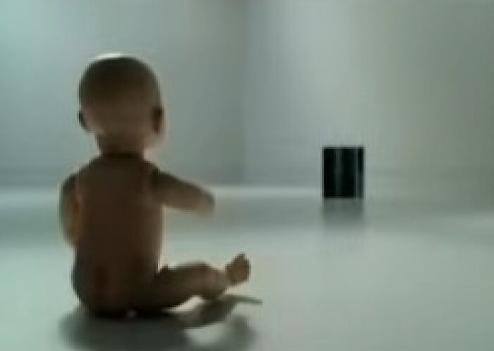 Baby doll sitting on a floor facing a black object in an empty room, evoking creepy commercial scenes.