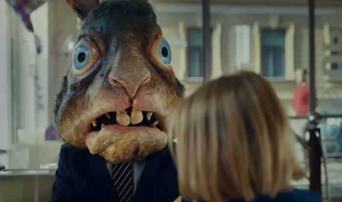 Person wearing a creepy large rabbit mask with wide eyes and crooked teeth in a disturbing commercial scene.