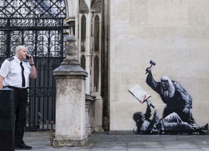 Security guard on phone near street art depicting chaotic good judge holding gavel over a fallen figure on wall.