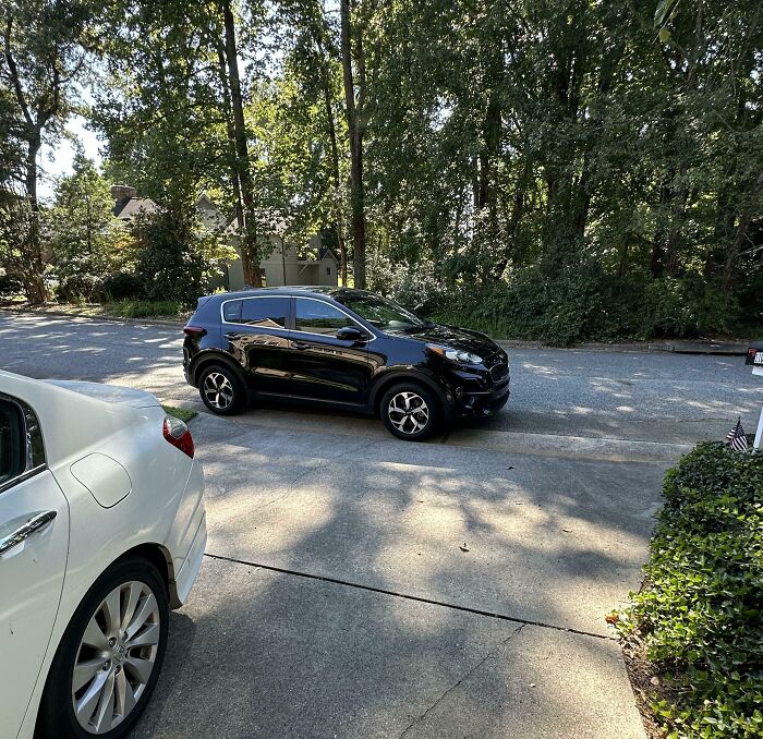 Black car parked across driveway blocking white car in suburban setting, illustrating people allergic to acting like sensible adults.
