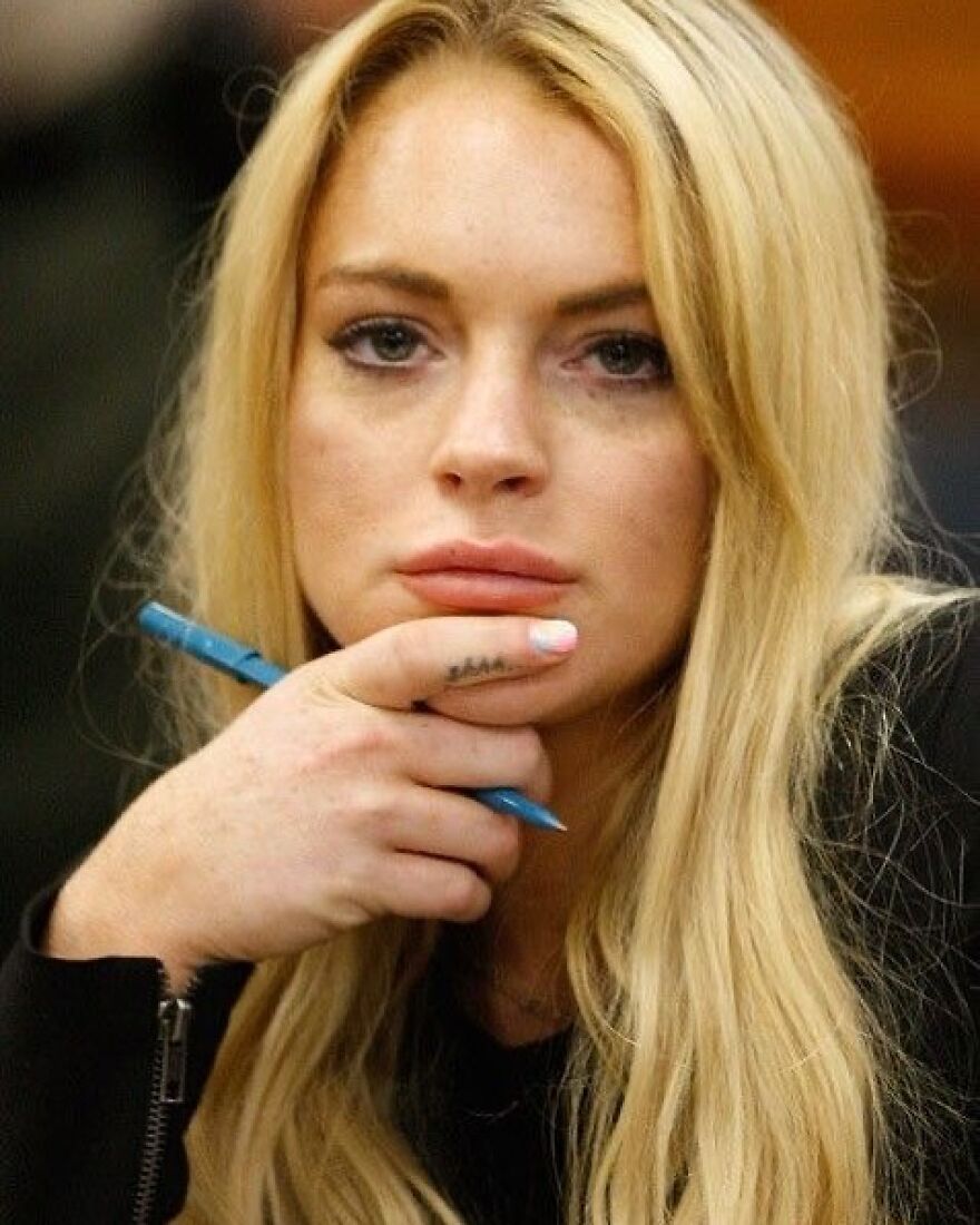 Lindsay Lohan with long blonde hair, holding a pen near her face, showing a contemplative expression in an indoor setting.