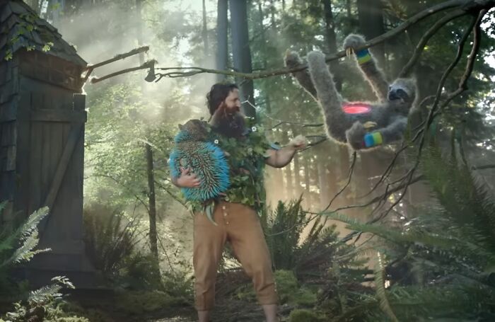 Man in a forest holding a quirky creature while a colorful sloth hangs from a tree in a creepy commercials scene.