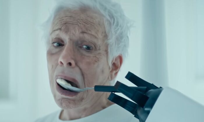 Elderly woman looking scared as a robotic hand feeds her with a spoon in a creepy commercial scene.