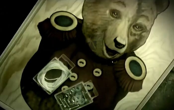 Vintage creepy bear doll lying in a box surrounded by buttons and string, evoking unsettling commercial imagery.