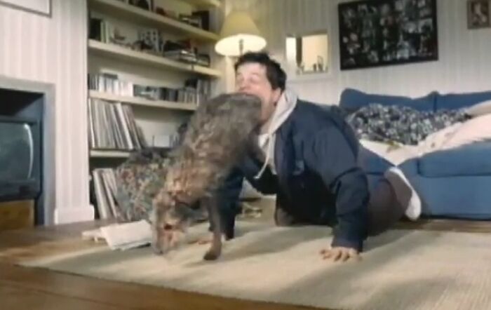 Man on hands and knees in a living room with a dog licking his face in a creepy commercial scene.